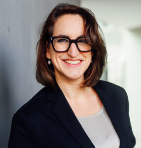 Das Bild zeigt ein fotografiertes Portrait von einer weißen Person mit schulterlangen braunen Haaren in einem Seitenscheitel gestylt, einer schwarzen großen Brille und einem großen Lächeln. An ihren Ohren trägt sie Perlenohrringe. Sie trägt ein graues T-Shirt und darüber ein schwarzes Jackett. Das Fpoto ist vor einem grauen Hintergrund aufgenommen, welcher schwach an ein Gebäude aus Beton erinnert.
